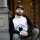 Man with a beard sitting outside, wearing a black cap and a white sweatshirt with logo, leaning against a pillar