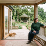 Person sitting on bench in rustic open-plan room, overlooking garden with gazebo and countryside, dog lying nearby